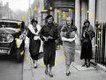 Vier Frauen in eleganter 30er Jahre Mode, Pelzmänteln und Hüten spazieren an einem Oldtimer entlang.