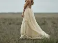 Braut in einem fließenden, cremefarbenen Kleid mit Tupfenmuster, das perfekt zum **dresscode boho hochzeit** passt. Sie steht in einer weiten, grasbewachsenen Ebene.