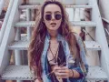 Frau mit langen Haaren und Sonnenbrille sitzt auf einer Metalltreppe. Sie trägt ein offenes Jeanshemd, das sie lässig mit einem Spitzen-Bralette kombiniert.