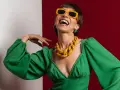 Lachende Frau in grünem Kleid mit auffälligem gelben Statement-Collier und Sonnenbrille, die aktuelle Trendfarben verkörpert.