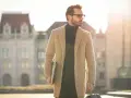 Ein Mann im old money style, mit Brille und beigem Mantel, trägt einen Laptop und eine Reisetasche.