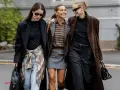 Drei Frauen in komischer Mode, die lachend nebeneinander gehen. Eine trägt einen braunen Lederblazer, die andere einen langen braunen Mantel.