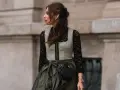 Eine Frau trägt eine graue Weste über einer schwarzen Spitzenbluse. Sie kombiniert die schwarze Bluse mit einem langen, olivgrünen Rock und einer kleinen schwarzen Tasche.