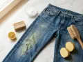 Blaue Jeans mit Grasflecken, Gallseife, Zitrone, Natron und Bürste zum Grasflecken entfernen.