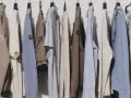 Eine Reihe von Kleidungsstücken, darunter Jacken, Pullover und Hosen, hängen an Kleiderbügeln. Diese zeitlose Kleidung in gedeckten Farben strahlt Komfort und Stil aus.