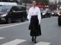 Eine Frau in einem weißen Hemd und einem schwarzen Plisseerock kombiniert mit Pumps und einer kleinen Tasche steht auf einem Zebrastreifen.