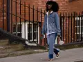 Frau in Jeans-Patchwork-Hose und Jeansjacke, ein Trend für den Jeans-Trend 2026. Weiße Sonnenbrille, weiße Sandalen.