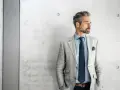 Stilvolles herren outfit sportlich elegant ab 50: Mann mit grauem Sakko, blauem Hemd und Krawatte vor Betonwand.