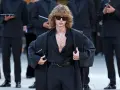 Ein Mann mit lockigem Haar und Sonnenbrille trägt eine schwarze, offen geknöpfte Bluse, weite Hosen und einen Mantel. Die Kleidung wirkt typisch 80er Jahre.