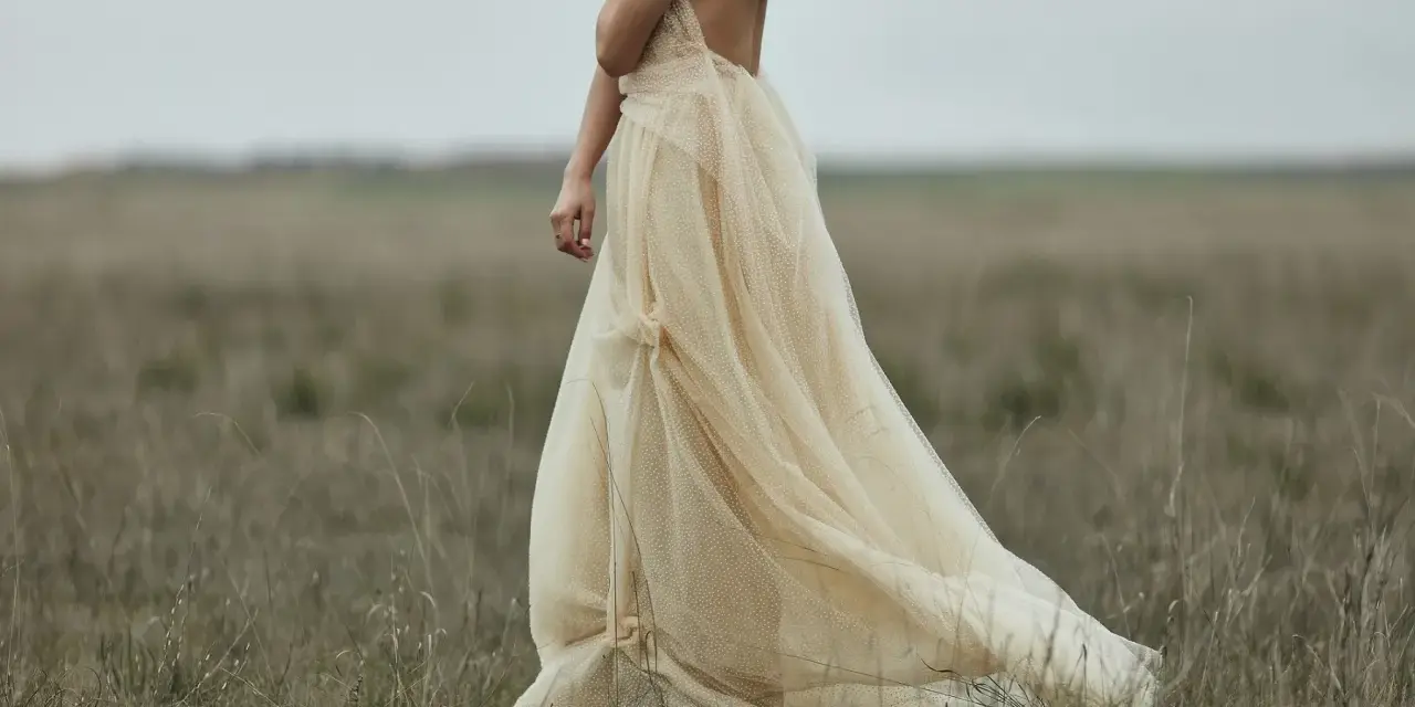 Braut in einem fließenden, cremefarbenen Kleid mit Tupfenmuster, das perfekt zum **dresscode boho hochzeit** passt. Sie steht in einer weiten, grasbewachsenen Ebene.