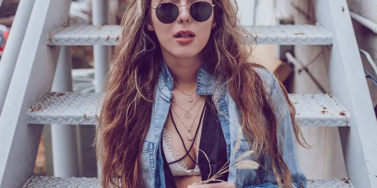 Frau mit langen Haaren und Sonnenbrille sitzt auf einer Metalltreppe. Sie trägt ein offenes Jeanshemd, das sie lässig mit einem Spitzen-Bralette kombiniert.