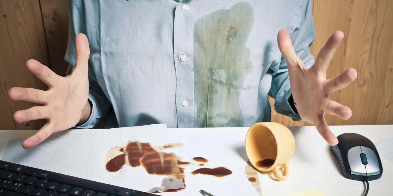 Mann mit verschüttetem Kaffee auf Hemd und Schreibtisch. Eingetrocknete Kaffeeflecken entfernen ist eine Herausforderung.