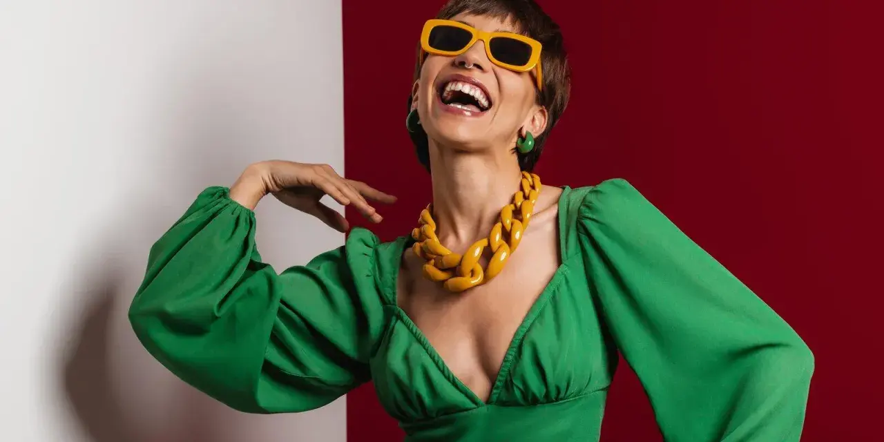 Lachende Frau in grünem Kleid mit auffälligem gelben Statement-Collier und Sonnenbrille, die aktuelle Trendfarben verkörpert.