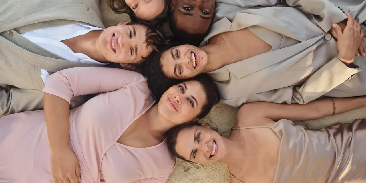 Sechs lächelnde Menschen liegen eng beieinander. Ihre Kleidung, wie ein beiger Anzug und ein rosafarbenes Kleid, inspiriert die Idee einer leichten basisgarderobe sommertyp.