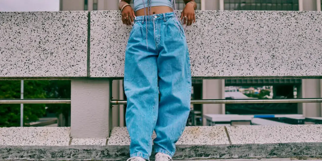 Person in einem lässigen baggy jeans outfit mit Crop-Top und weißen Sneakern steht auf einer Betonmauer.
