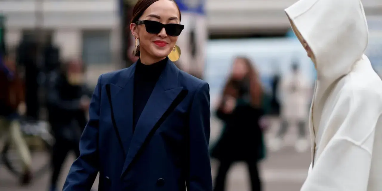 Eine Frau mit Sonnenbrille und auffälligem Schmuck trägt einen dunkelblauen Blazer über einem schwarzen Rollkragenpullover. Sie kombiniert dunkelblau und schwarz gekonnt.