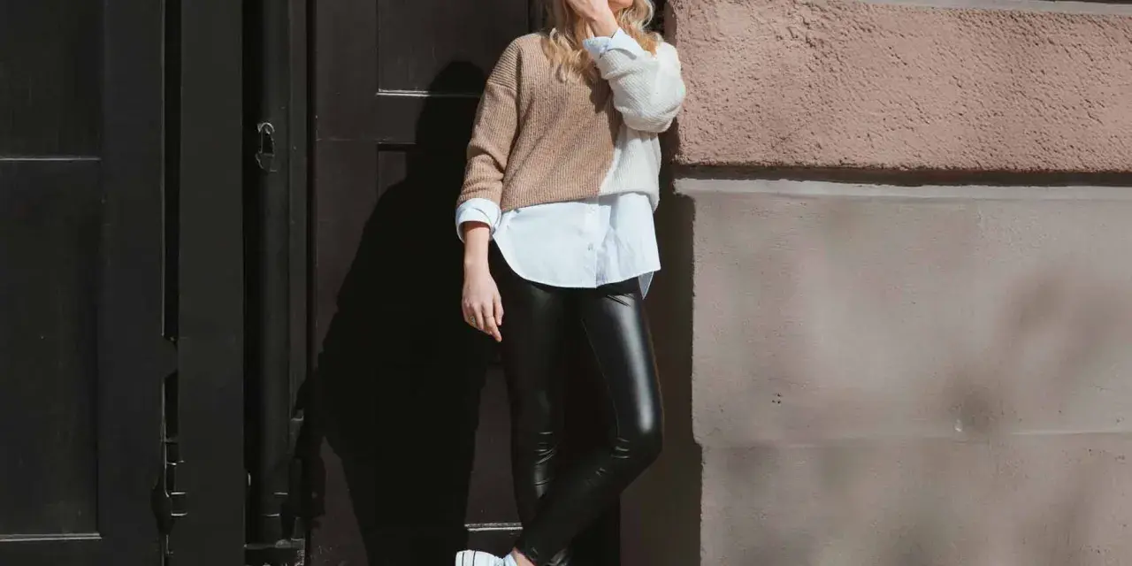 Eine Frau kombiniert elegant schwarze Hose mit einem zweifarbigen Pullover und weißem Hemd. Weiße Plateau-Sneaker runden den Look ab.