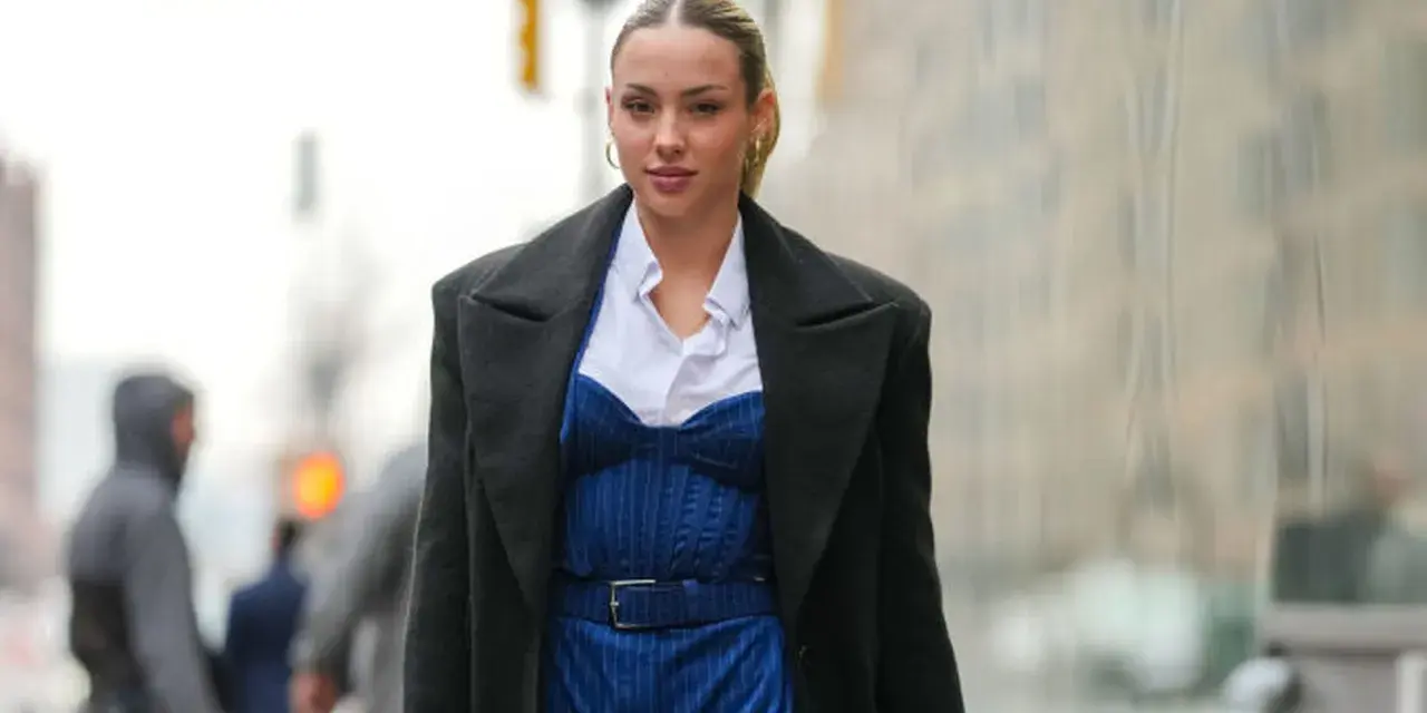 Frau trägt eine schwarze Jacke über einem blauen Nadelstreifen-Jumpsuit mit Korsett-Oberteil und weißem Hemd.