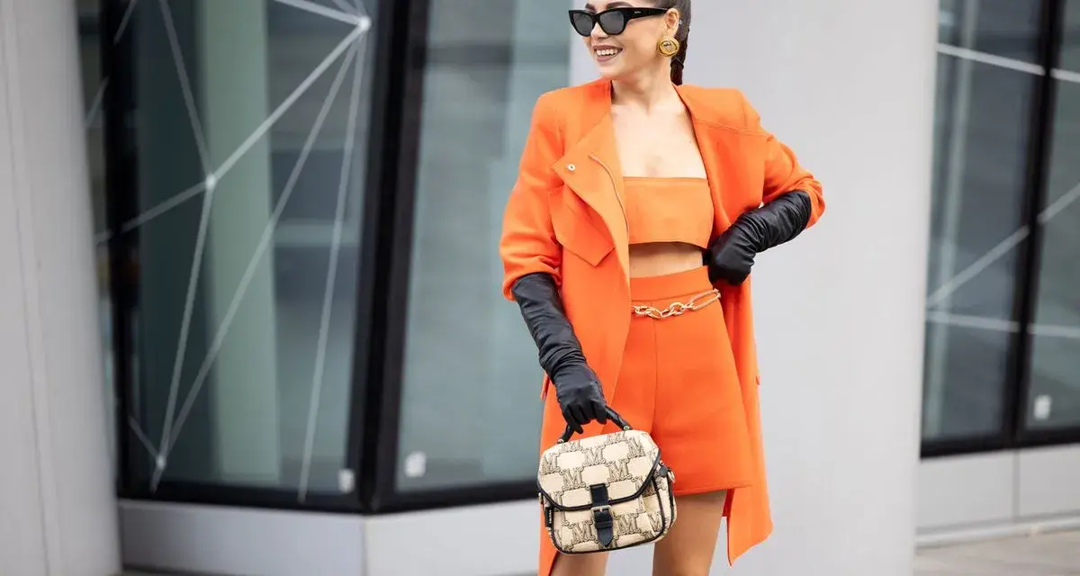 Eine Frau in einem leuchtend orangen Outfit mit schwarzen Handschuhen und einer Tasche. Schwarz passt wunderbar zu Orange, eine tolle Farbkombination!