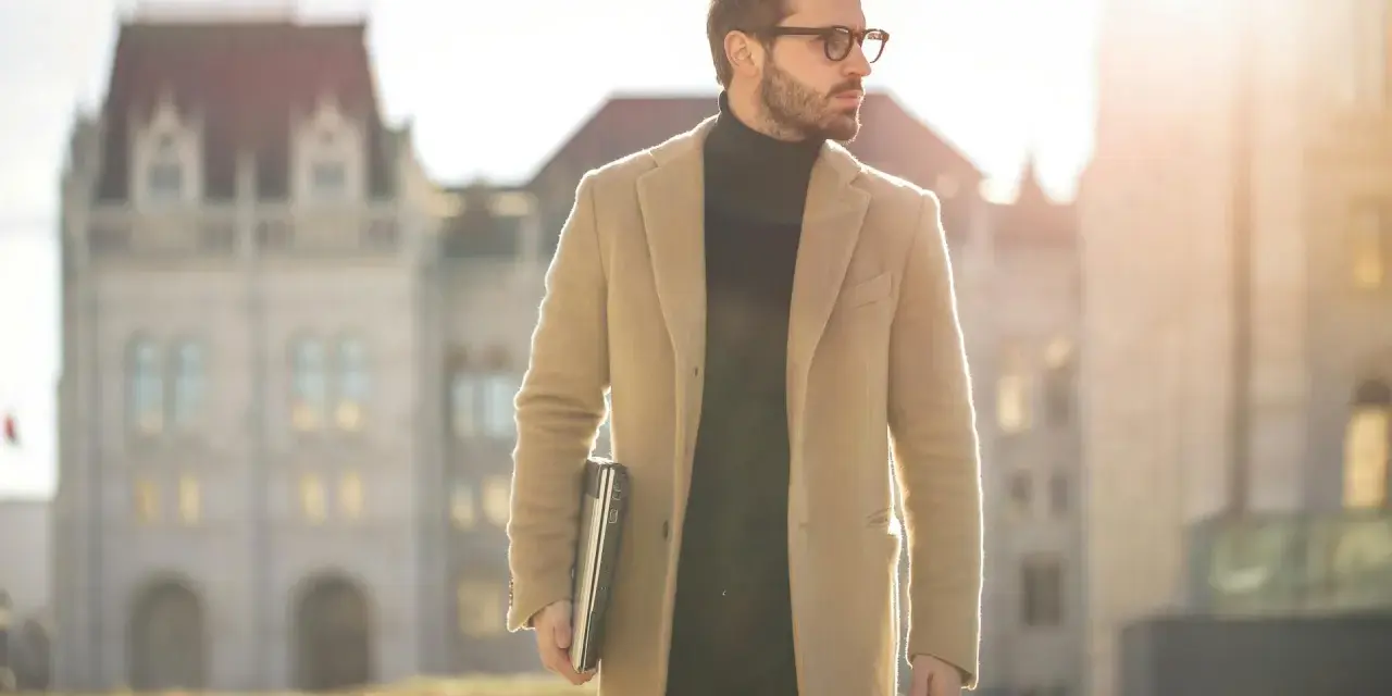 Ein Mann im old money style, mit Brille und beigem Mantel, trägt einen Laptop und eine Reisetasche.