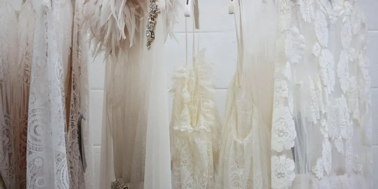 Mehrere zarte, weiße Brautkleider hängen an Kleiderbügeln. Eines hat eine Federverzierung. Die Reinigung eines Hochzeitskleides ist wichtig, damit es schön bleibt.
