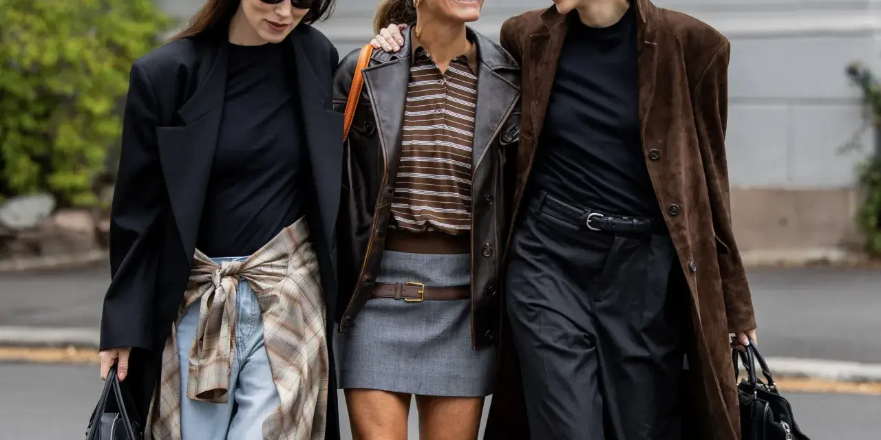 Drei Frauen in komischer Mode, die lachend nebeneinander gehen. Eine trägt einen braunen Lederblazer, die andere einen langen braunen Mantel.