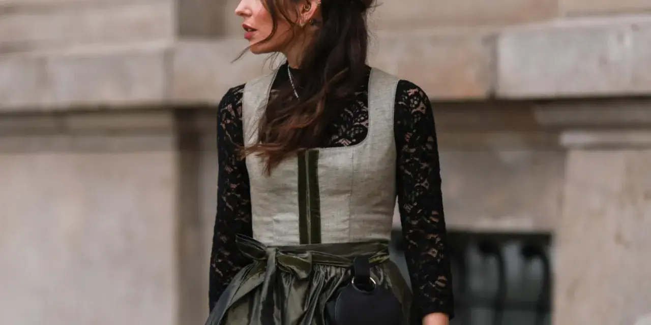 Eine Frau trägt eine graue Weste über einer schwarzen Spitzenbluse. Sie kombiniert die schwarze Bluse mit einem langen, olivgrünen Rock und einer kleinen schwarzen Tasche.