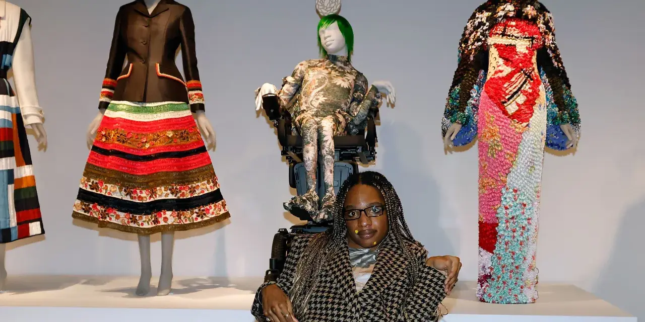 Bei einer Vernissage werden extravagante Kleider auf Schaufensterpuppen präsentiert. Eine Frau im Rollstuhl sitzt davor.