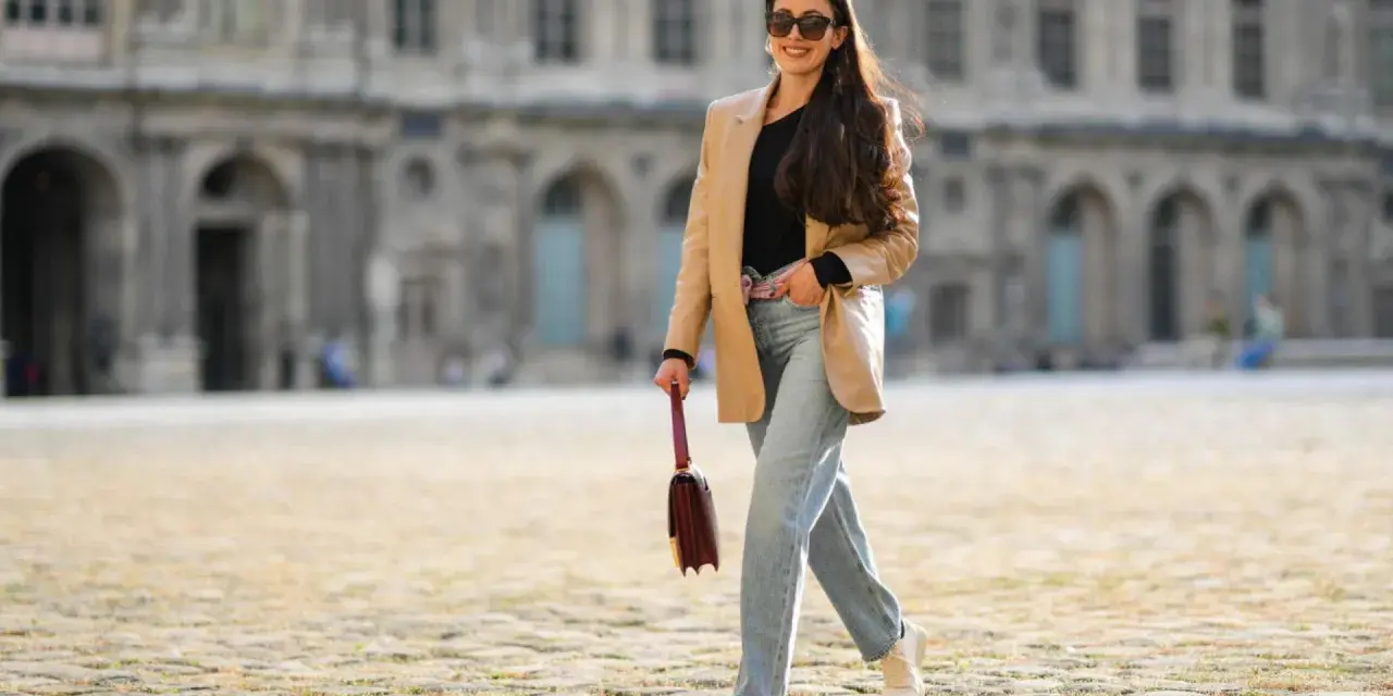 Frau in weiter Jeans und blauem Hemd mit goldener Clutch und Glitzer-Schuhen überquert Straße.