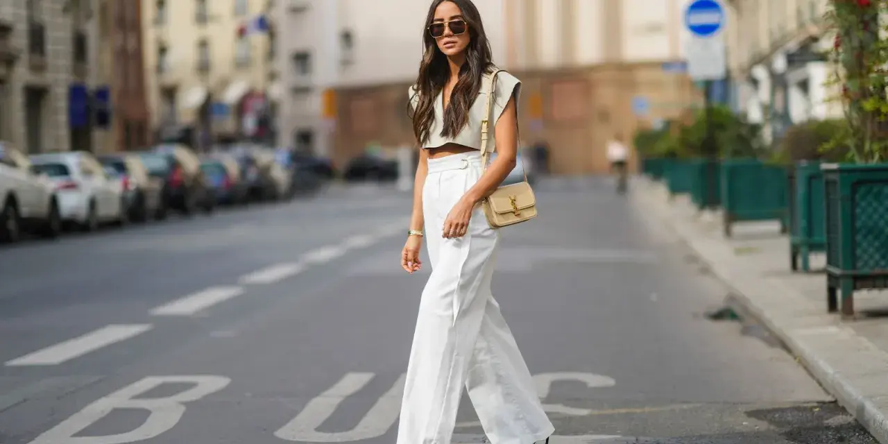 Stilvolle Dame in Paris, die eine weiße Leinenhose kombiniert mit einem kurzen Top und einer Umhängetasche trägt.
