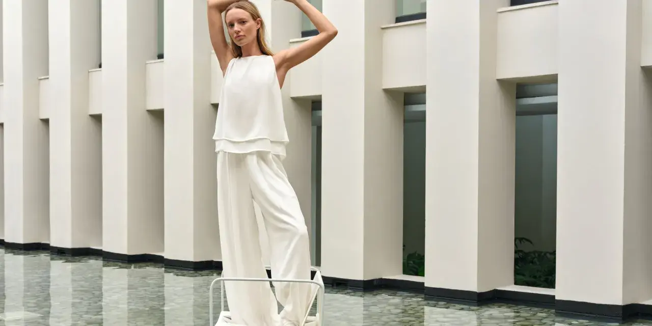 Frau in weißem Outfit mit weiten Palazzo Hosen, elegant gestylt vor moderner Architektur.