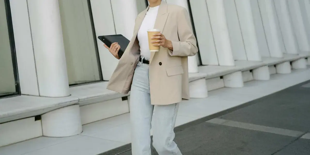 Frau im smart casual Dresscode mit Blazer, Jeans und Kaffee. Sie hält ein Tablet.