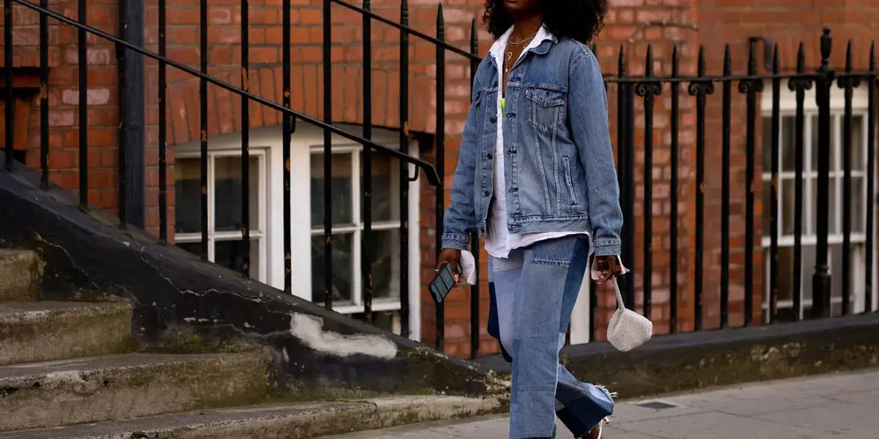 Frau in Jeans-Patchwork-Hose und Jeansjacke, ein Trend für den Jeans-Trend 2026. Weiße Sonnenbrille, weiße Sandalen.