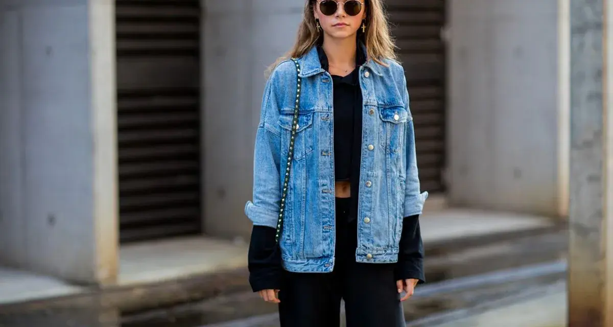 Stylische Frau mit lässiger Jeansjacke kombinieren mit schwarzem Crop-Top und Hose. Sonnenbrille und kleine Tasche runden den Look ab.