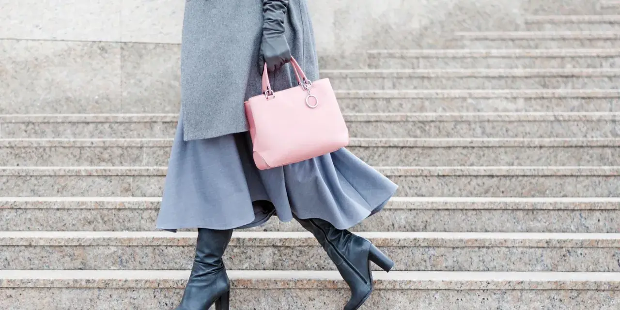 Stilvoll **Kleider im Winter tragen**: Grauer Mantel, blauer Rock, schwarze Stiefel und eine rosafarbene Tasche auf Steinstufen.