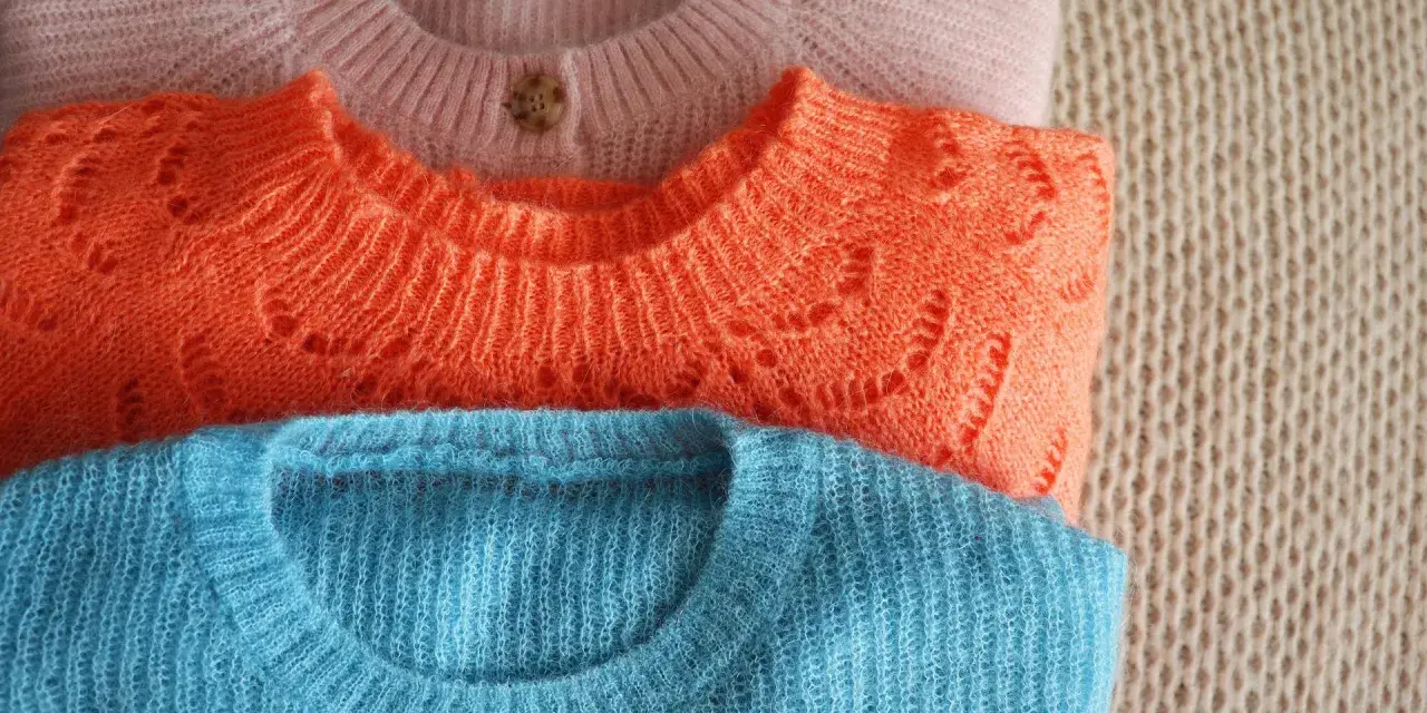 Gestapelte Pullover in Rosa, Orange und Blau. So kannst du deine **Wolle richtig waschen** und sie bleibt schön.