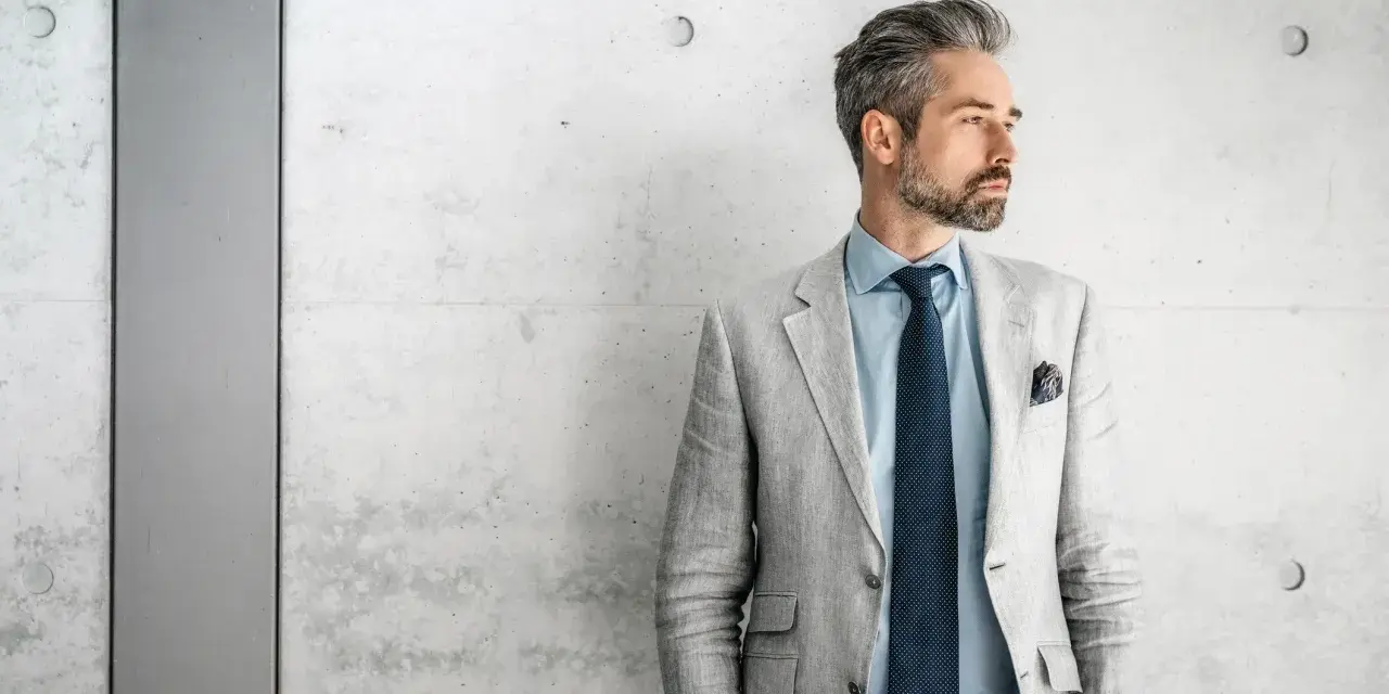 Stilvolles herren outfit sportlich elegant ab 50: Mann mit grauem Sakko, blauem Hemd und Krawatte vor Betonwand.
