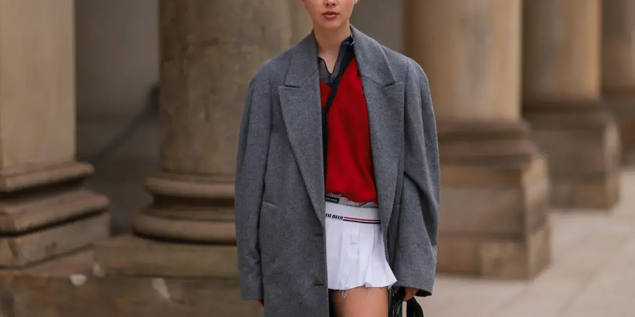 Ein junges Model präsentiert einen preppy look mit grauem Mantel, rotem Pullover und weißem Faltenrock.