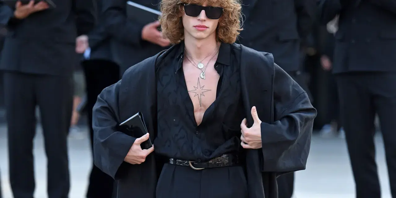 Ein Mann mit lockigem Haar und Sonnenbrille trägt eine schwarze, offen geknöpfte Bluse, weite Hosen und einen Mantel. Die Kleidung wirkt typisch 80er Jahre.