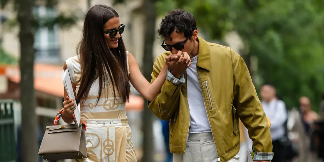 Ein Mann küsst die Hand einer Frau. Ihr **outfit erstes date** ist elegant: Sie trägt ein gemustertes Kleid, er eine olivgrüne Jacke.