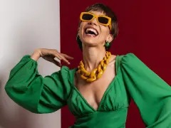 Lachende Frau in grünem Kleid mit auffälligem gelben Statement-Collier und Sonnenbrille, die aktuelle Trendfarben verkörpert.