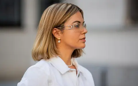 Frau mit einem stylischen **long bob** und auffälligen Sonnenbrillen, trägt eine weiße Jacke und Perlenschmuck.