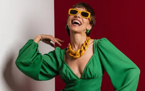 Lachende Frau in grünem Kleid mit auffälligem gelben Statement-Collier und Sonnenbrille, die aktuelle Trendfarben verkörpert.