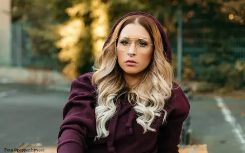 Frau mit blonden Haaren und Brille trägt einen Kapuzenpullover in dunklem Beere. Die Farbe Beere kombiniert sie gekonnt mit ihrem Outfit.