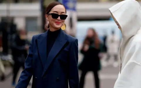 Eine Frau mit Sonnenbrille und auffälligem Schmuck trägt einen dunkelblauen Blazer über einem schwarzen Rollkragenpullover. Sie kombiniert dunkelblau und schwarz gekonnt.