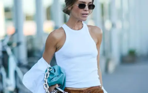 Frau im weißen Ripptop und Lederhose, trägt eine blaue Tasche mit silberner Kette. Der "ohne BH"-Trend ist hier subtil umgesetzt.