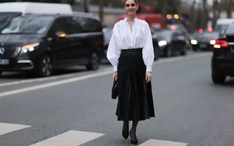 Eine Frau in einem weißen Hemd und einem schwarzen Plisseerock kombiniert mit Pumps und einer kleinen Tasche steht auf einem Zebrastreifen.