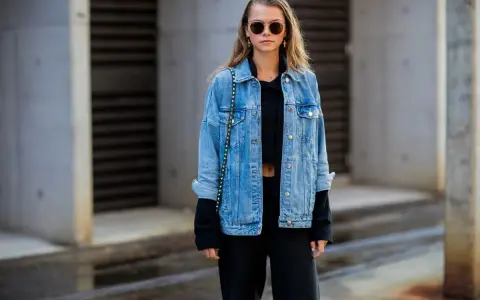 Stylische Frau mit lässiger Jeansjacke kombinieren mit schwarzem Crop-Top und Hose. Sonnenbrille und kleine Tasche runden den Look ab.