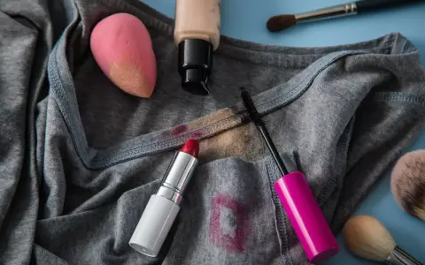 Lippenstiftflecken entfernen: Roter Lippenstift, Mascara, Foundation und Make-up-Schwamm auf grauer Kleidung.