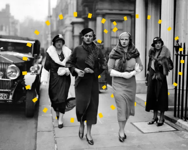Vier Frauen in eleganter 30er Jahre Mode, Pelzmänteln und Hüten spazieren an einem Oldtimer entlang.
